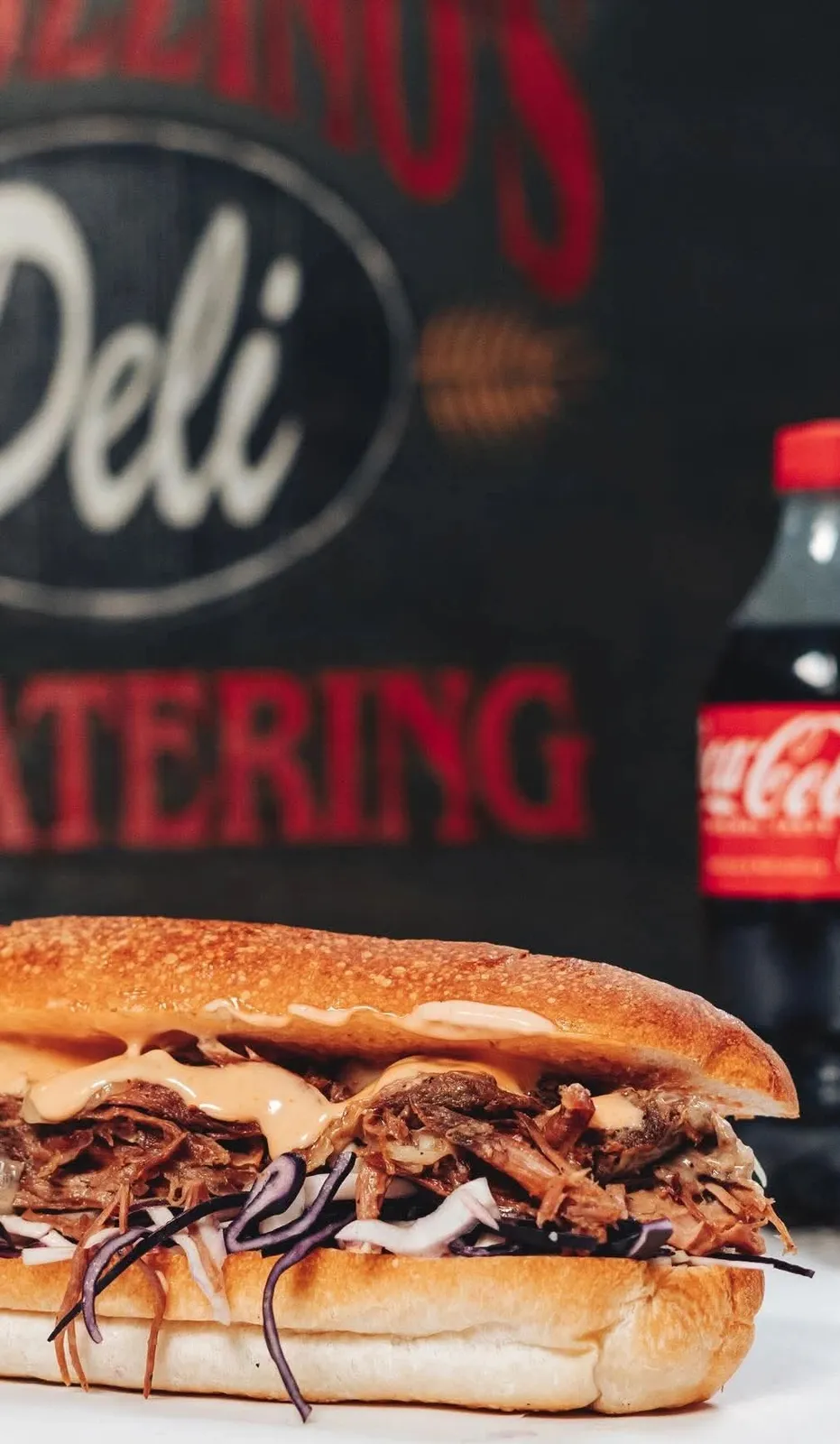 A pulled pork grinder with slaw and a bottle of Coke in front of the Ragozzino's Deli sign